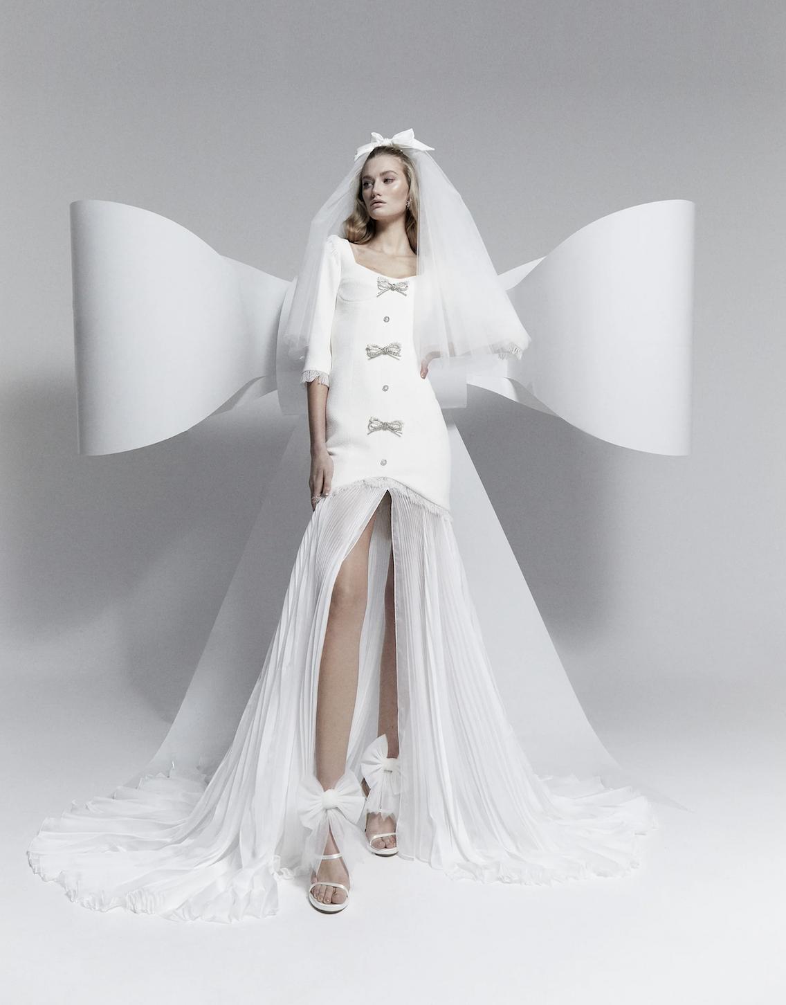 bride standing in front of a huge bow, wearing a bow and veil in hair, and a short wedding dress with glittery bow details and long train skirt underneath