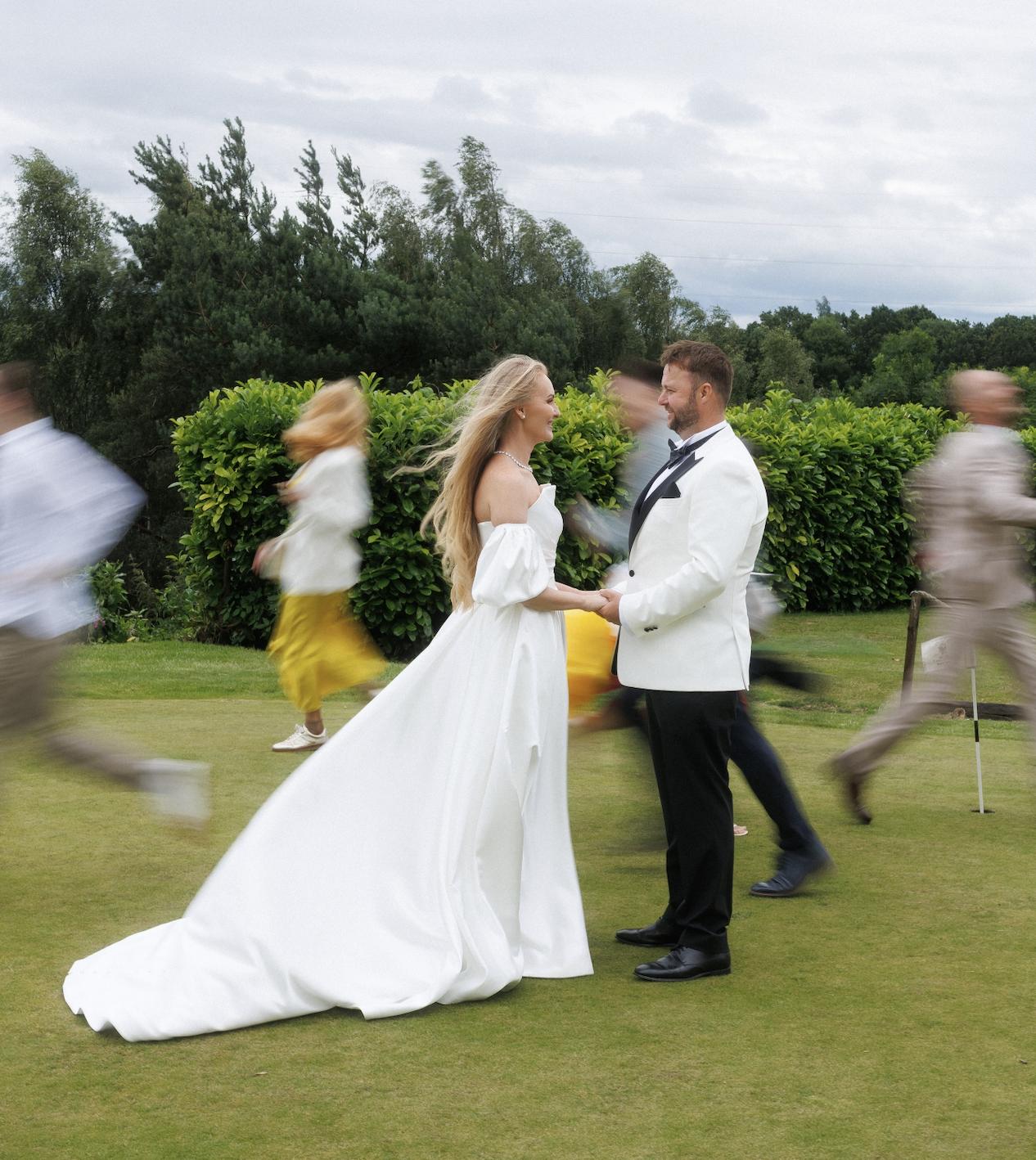 a bride and groom holding hands on a lawn whilst other guests blur into the background