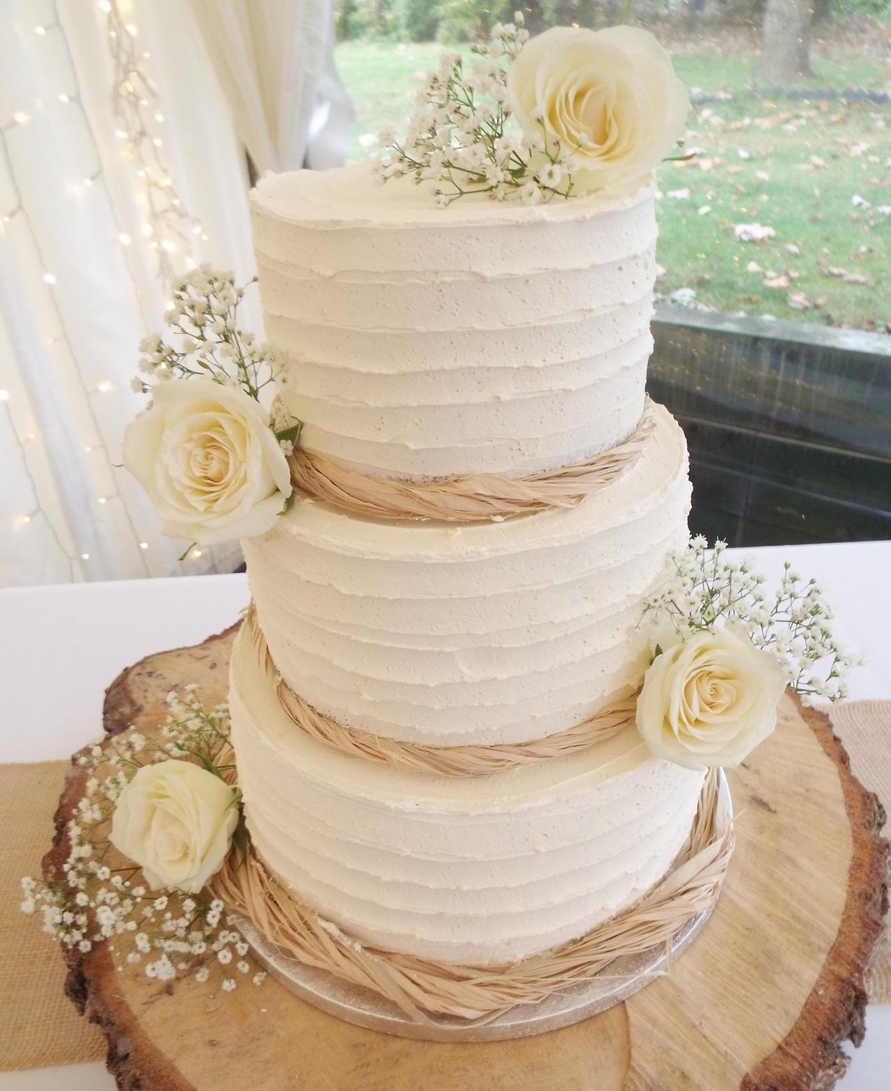 A three tier buttercream wedding cake with raffia detail and white flowers on a log slice.