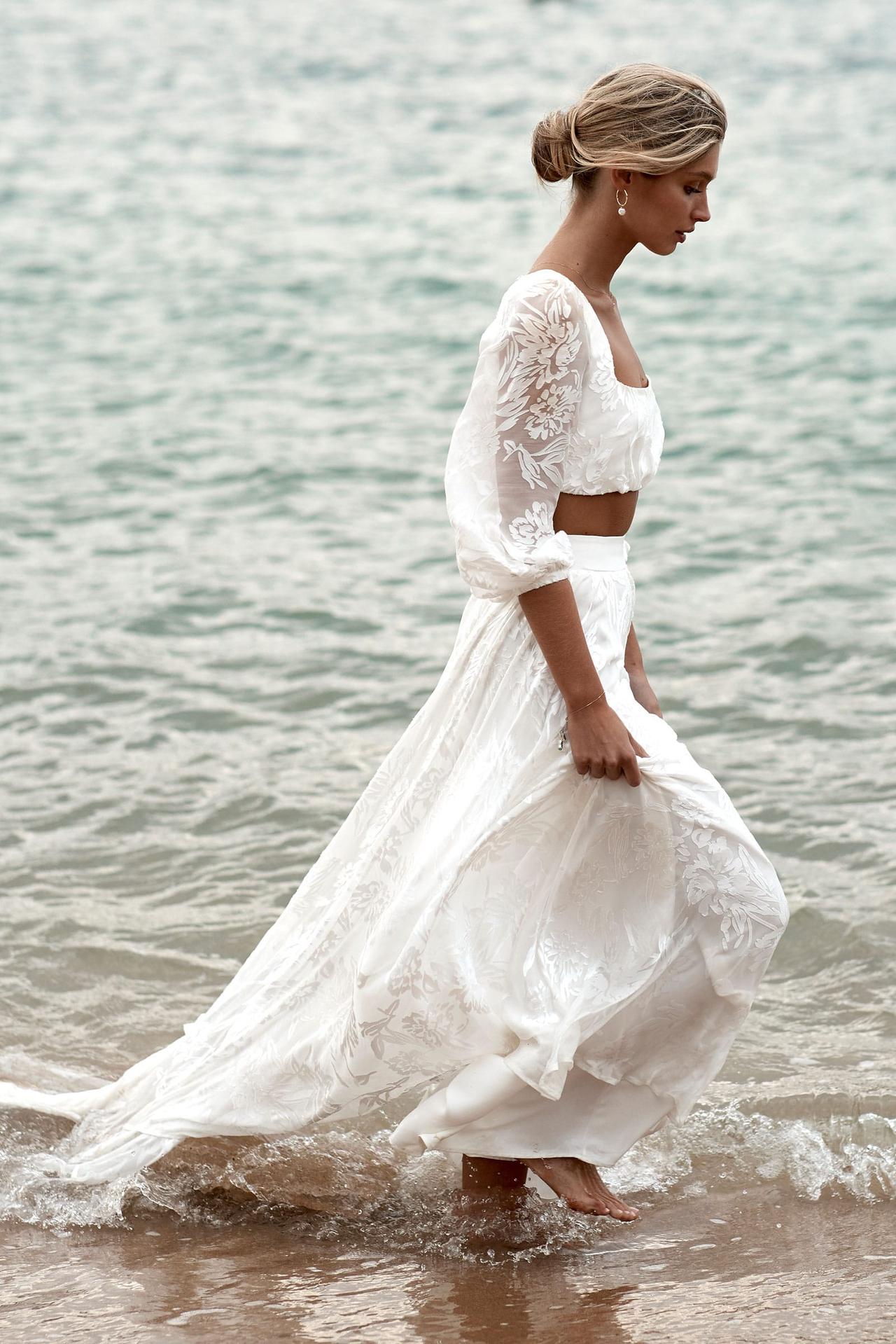 woman walking through gentle waves at the edge of the sea, wearing a two-piece bridal outfit with long skirt and long puff sleeve top