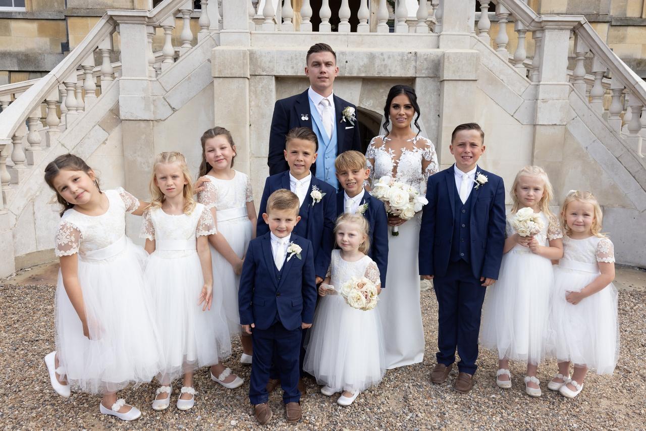 a bride and groom pose with four page boys in navy suits and eight flower girls in white lace flower girl dresses