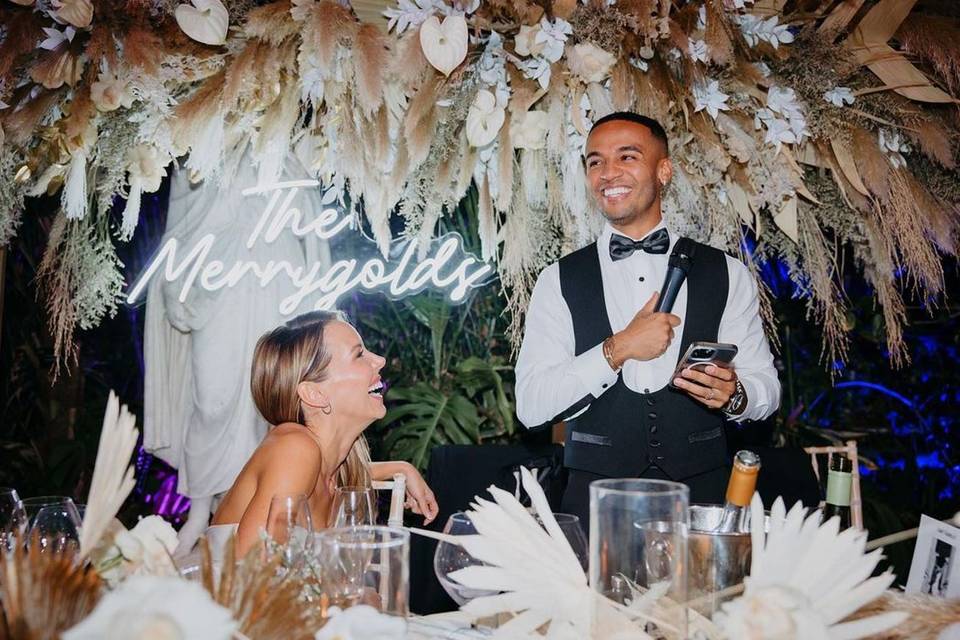 Aston and Steph Merrygold at their wedding. Aston is stood up making a speech while his bride watches him laughing and a sign saying 'The Merrygolds' is behind them