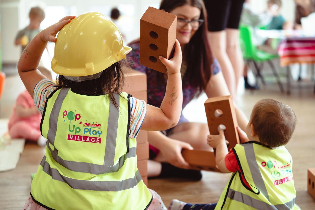 A toddler and a baby playing with foam bricks at a play village