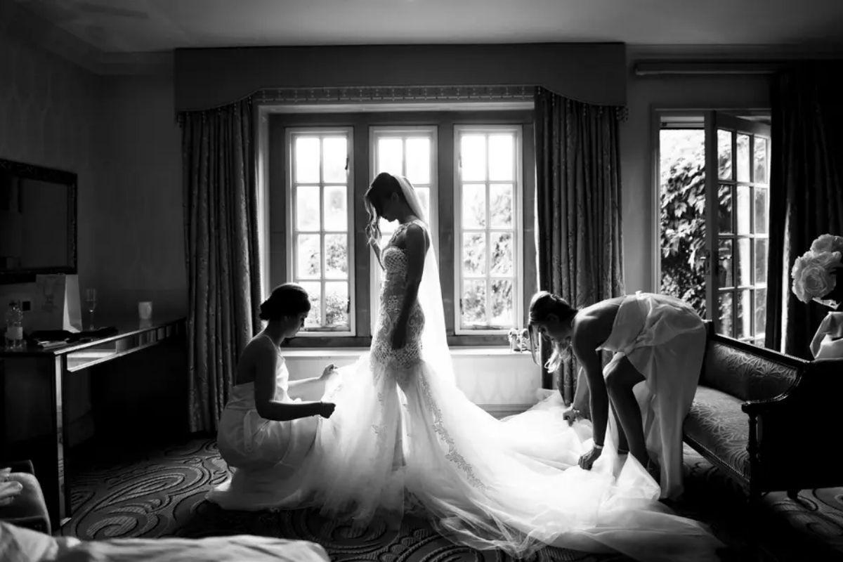 a bride wearing her wedding dress in a black and white shot as her bridesmaids position her train helping her get ready