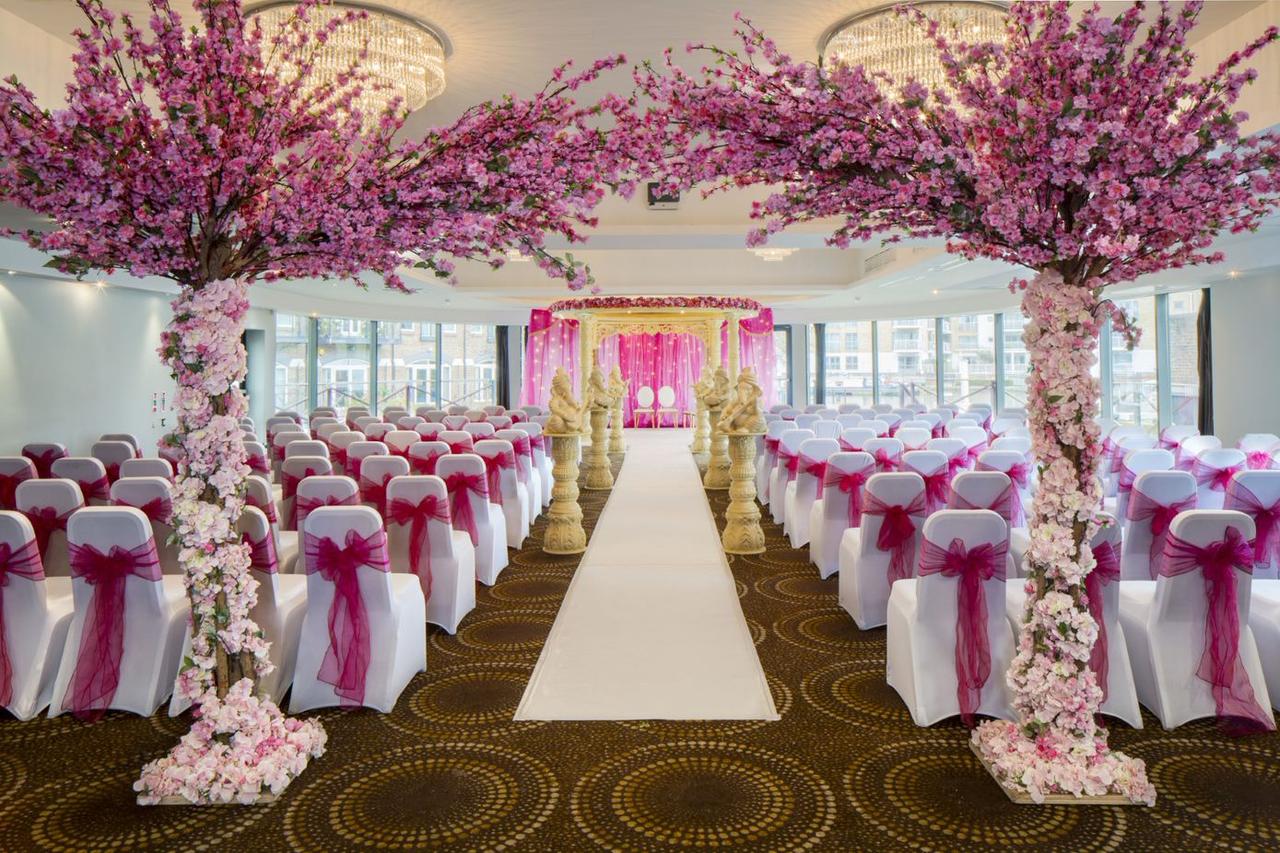 Interior of a hotel function room set up for a ceremony with white chair covers and hot pink sashes. There are two pink blossom trees in the foreground.