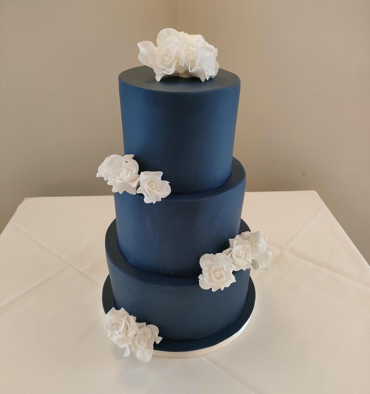 A dark blue iced fondant three tier cake with white sugar roses.