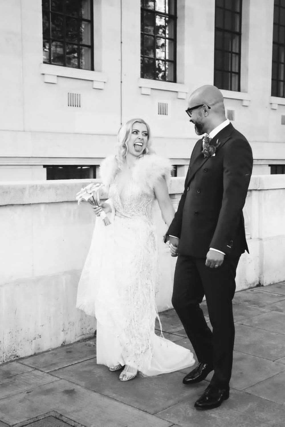 Black and white photo of a newlywed couple with a white bride with blonde hair sticking her tongue out at her groom wearing an embellished v-necked wedding dress with a white fur cape and simple bouquet 