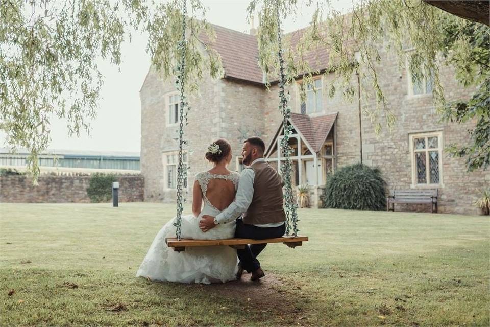 Bride and groom sit on a swing hanging from a tree outside a grey stone wedding venue