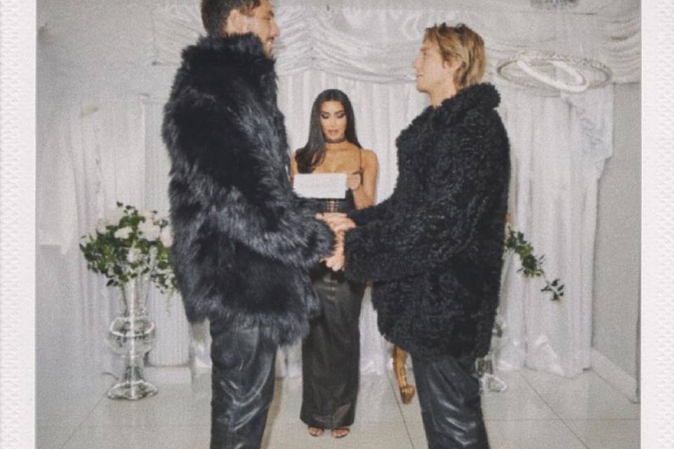 Kim Kardashian wears a black leather dress reading from paper as two men in black fur coats old hands at the altar