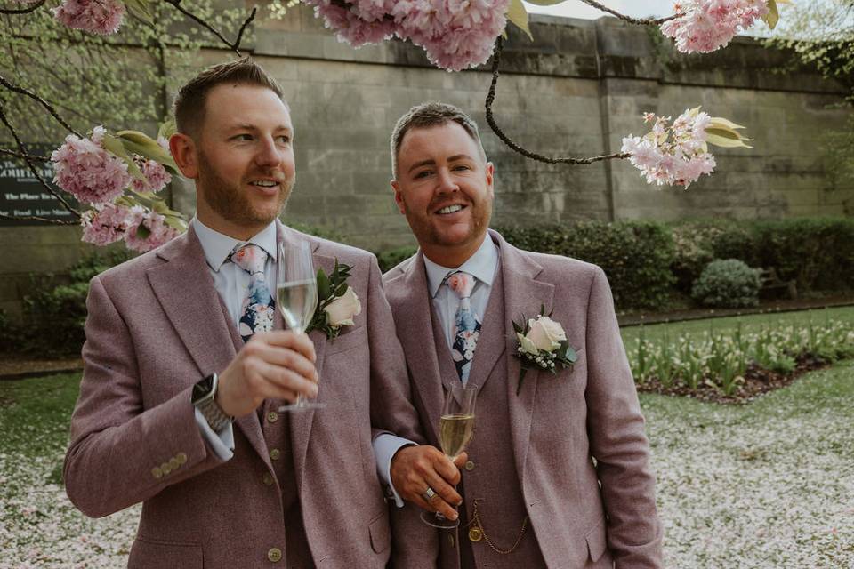 two grooms wear light pink tweed suits and drink champagne underneath a blossom tree