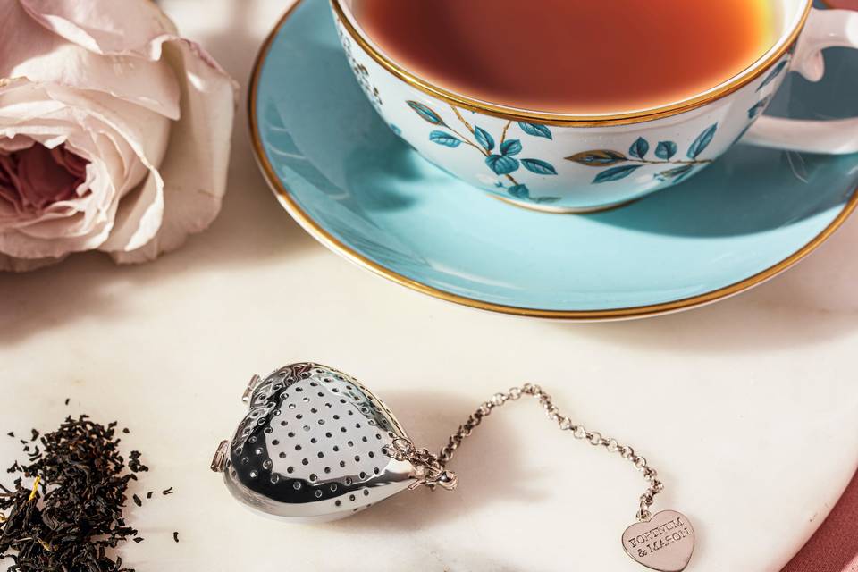A silver heart-shaped tea strainer next to a cup of tea and some loose leaf tea.