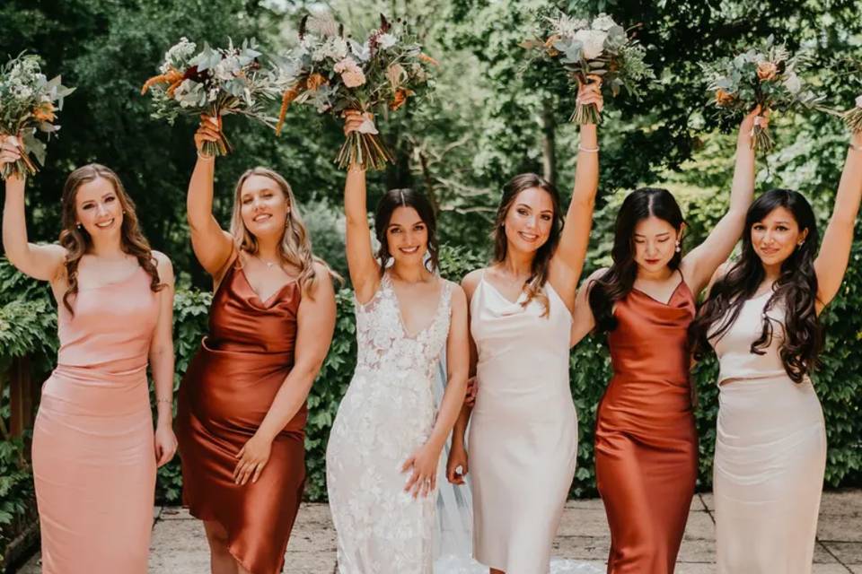 A bride and her five bridesmaids all holding their bouquets up in the air. All the bridesmaids are wearing mix and match bridesmaid dresses in nudes, oranges and rust colours 