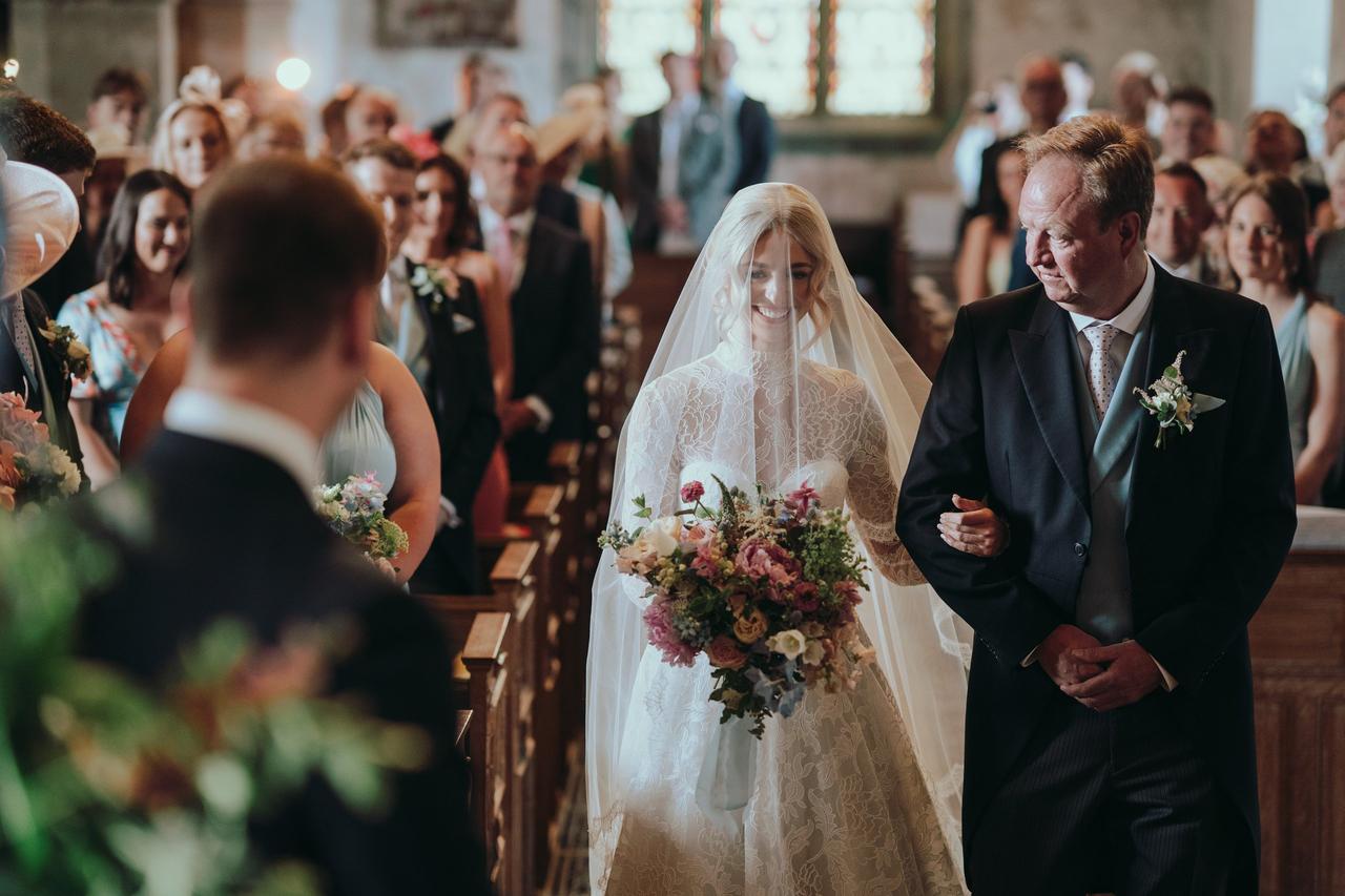 A bride walks down the aisle with her father in a church as guests look on.
