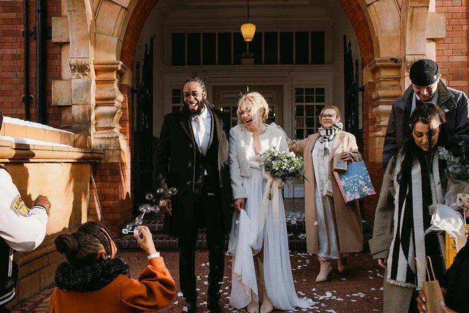 Bride and groom exit a venue laughing while their guests throw confetti and blow bubbles