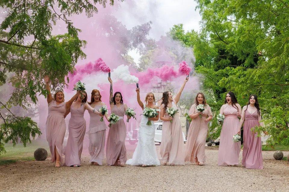 A line up of eight bridesmaids all in floor length pink bridesmaid dresses, walking along with the bride in the middle. The bride holds a white smoke cannon aloft whilst the bridesmaids hold up pink ones