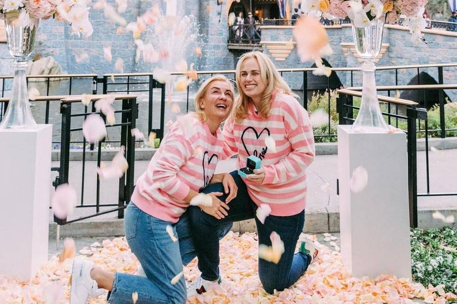 rebel wilson and her girlfriend announce engagement nextto two pillars at disney with petals everywhere