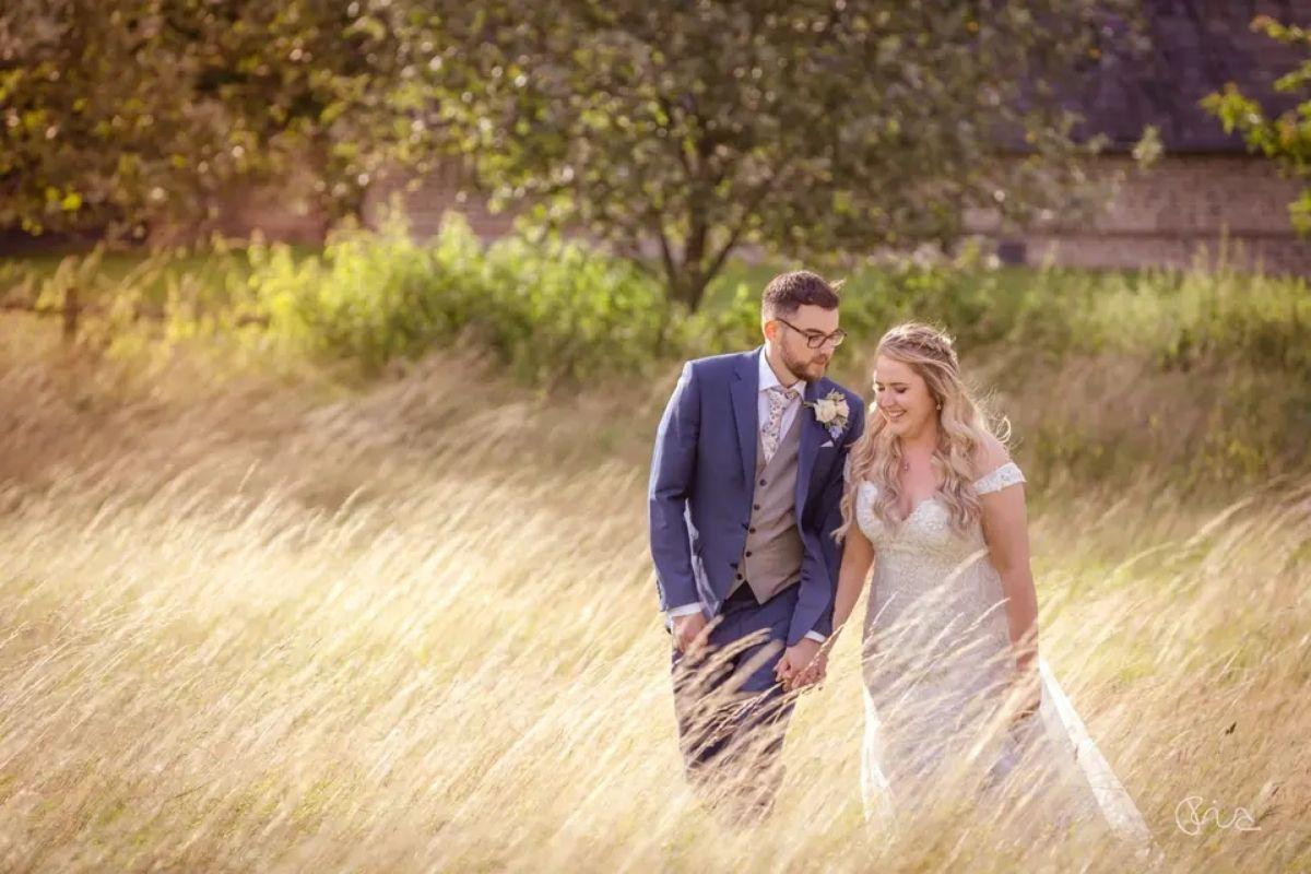 a bride and groom pose as they walk through a countryside field with long beige grass