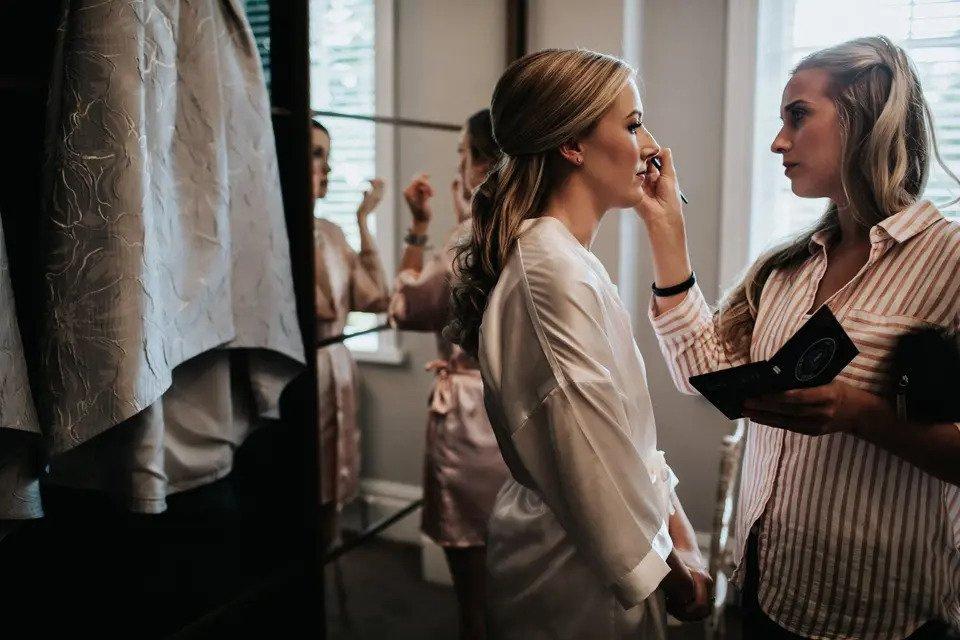 A bride stands in front of a mirror while her makeup is adjusted by an MUA