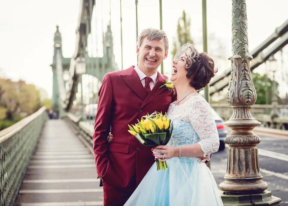 Smiling bride and groom embrace each other on Hammersmith Bridge with a bouquet of yellow flowers