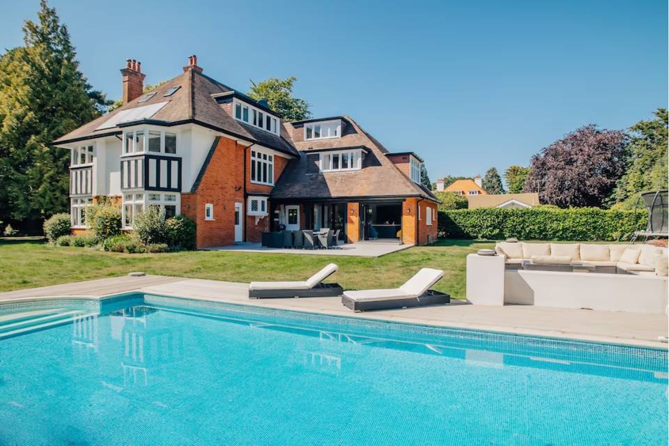 A swimming pool with sun loungers and a lawn behind it in front of a large hen party house