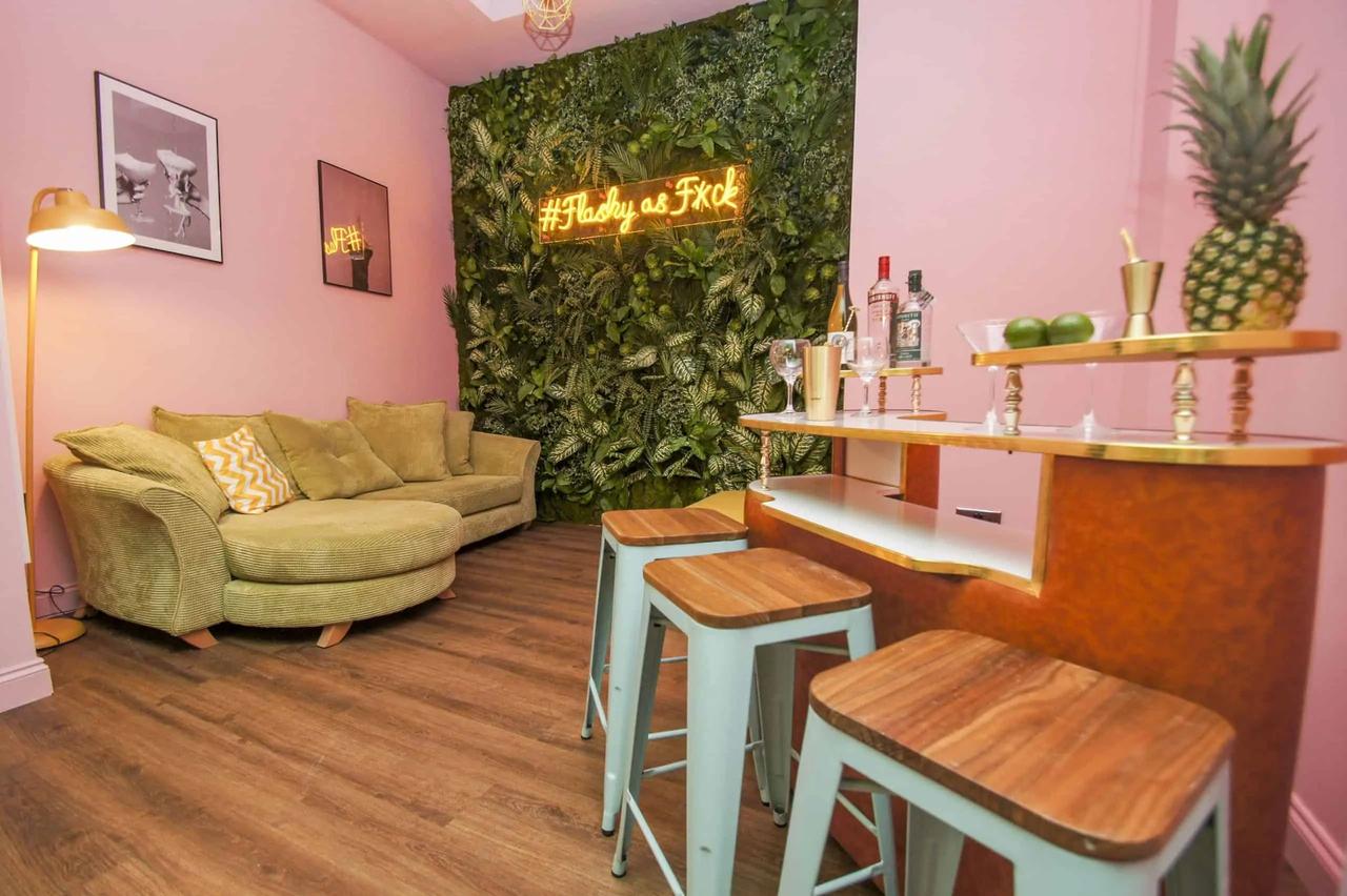 A pink room furnished with a vintage bar holding glasses and drinks, next to a yellow plush sofa and a gold lamp. The furthest wall is covered in green leaves, with a yellow light up sign hung in front