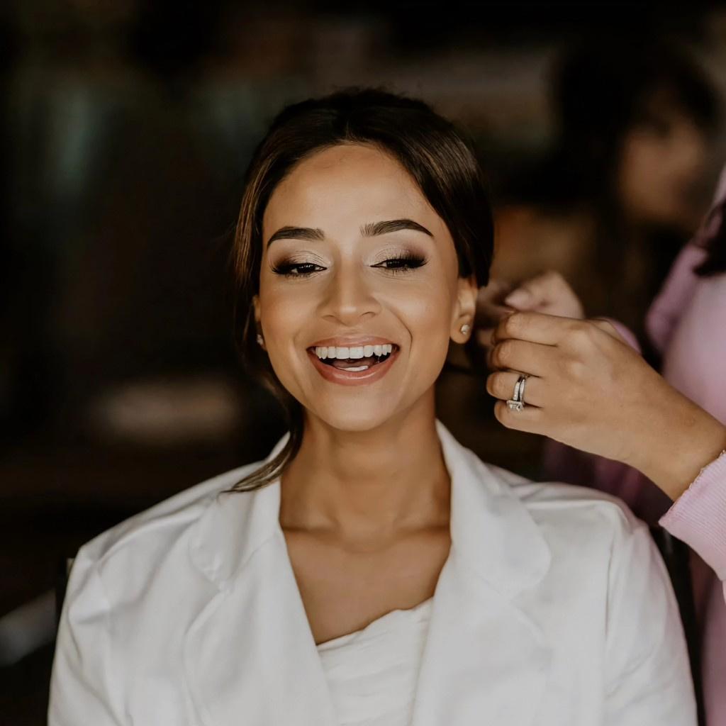 An Indian bride in a white robe laughing as she gets her makeup and hair done for her wedding day