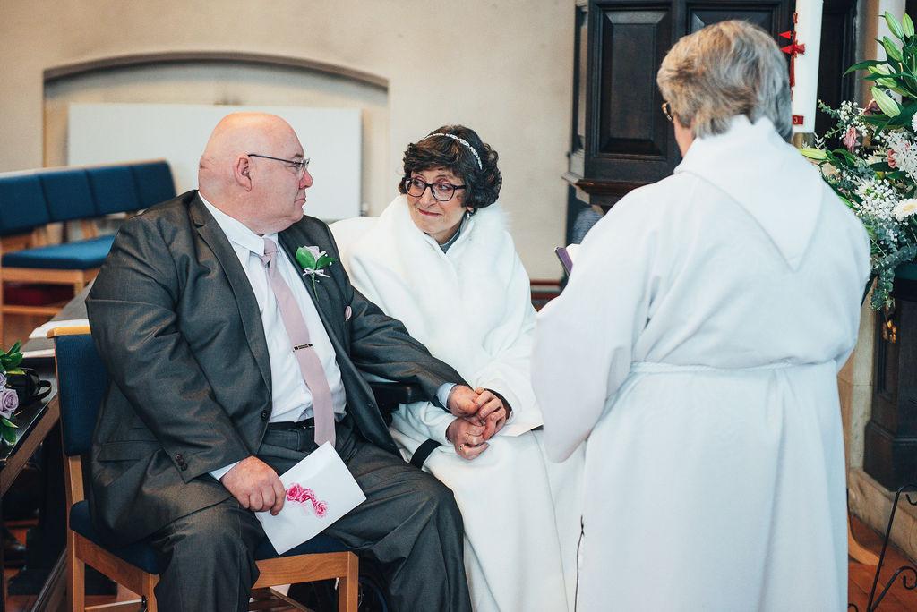A bride in a wedding dress in a wheelchair with her guide dog on one side of her and her groom sitting on the other side holding her hand