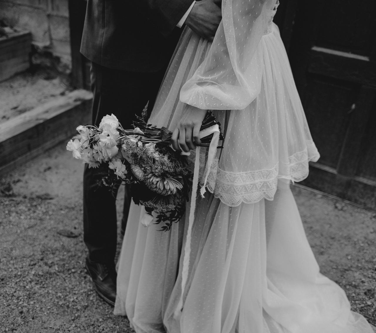 Black and white photo of the bottom half of a groom hugging a bride who is wearing a sheer, dotted multi-layered vintage style wedding dress and holding a wild bouquet