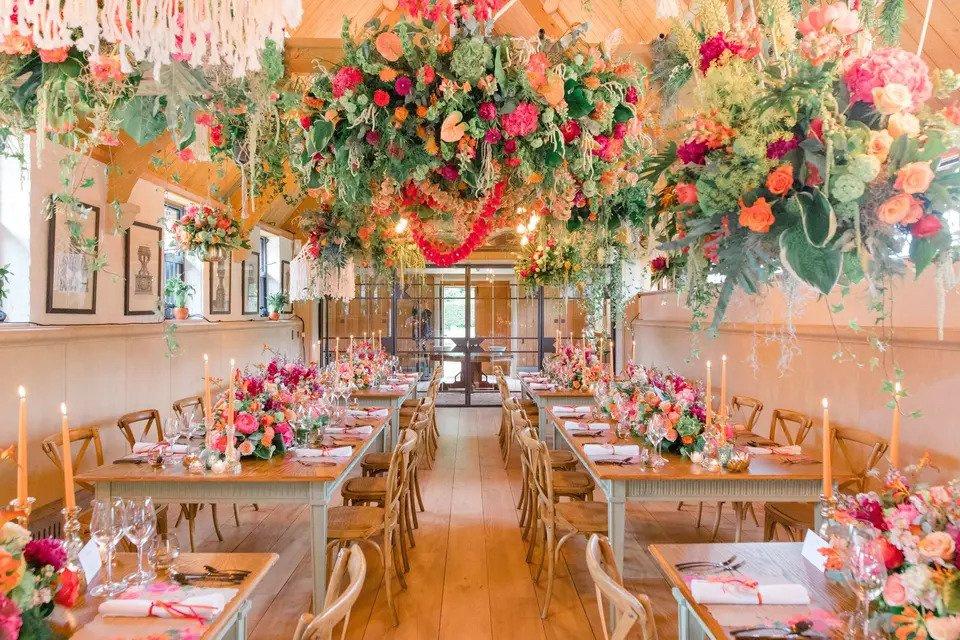 Bright room set for a wedding meal with long wooden tables, wooden chairs, glass panelled room dividers, dinner candles and a bright organise, pink and green floral canapy and centrepieces