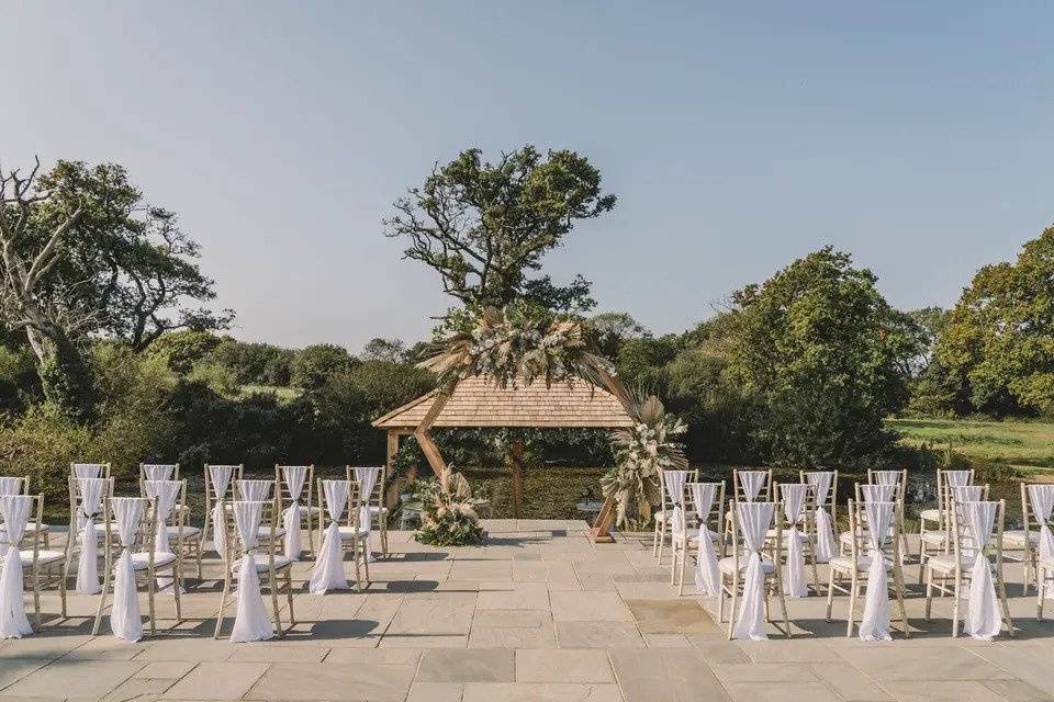 Outdoor wedding setup with wooden chairs and pergola surrounded by lawns and trees
