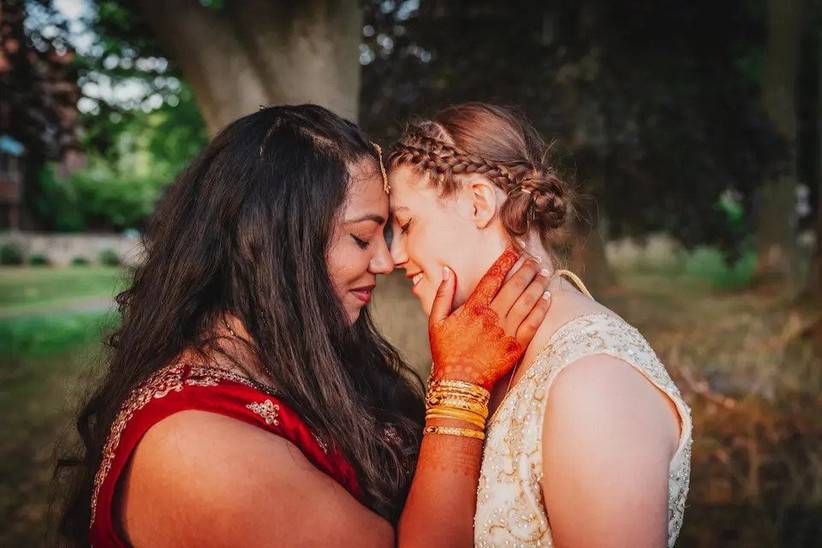Two brides - one in traditional Indian bridal attire, one in a western style white wedding dress - touching their foreheads together
