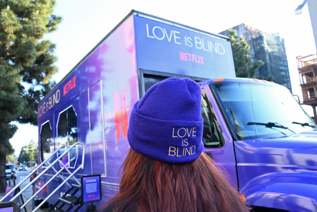 a woman wearing a love is blind hat walking towards a purple netflix bus outdoors