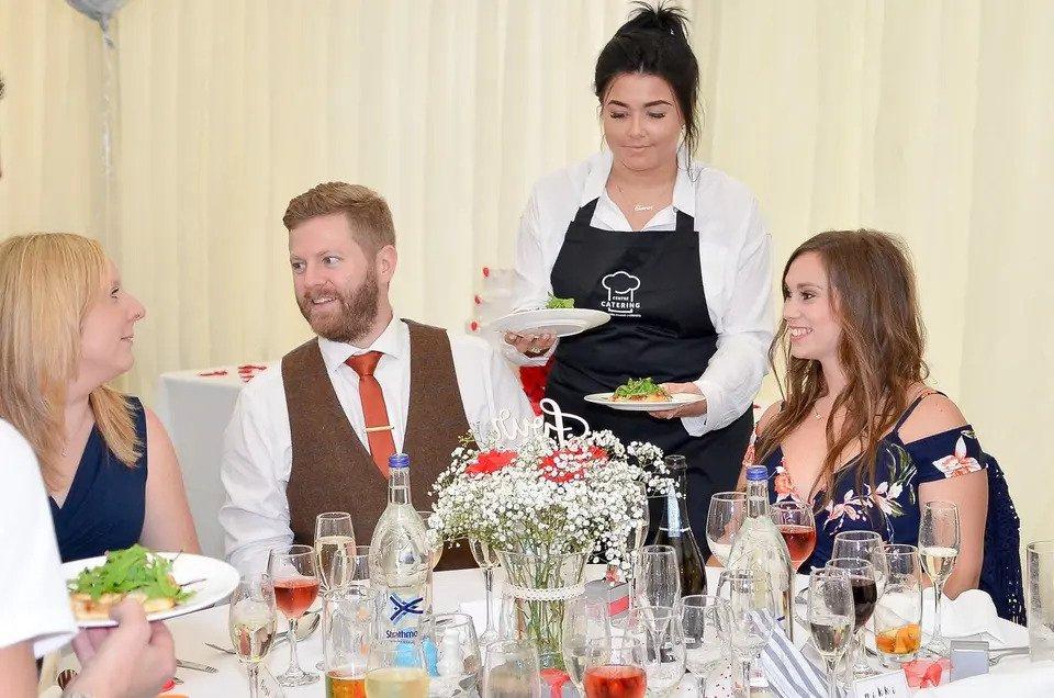 A waitress wearing a black Centre Catering apron serves three seated guests at a wedding meal
