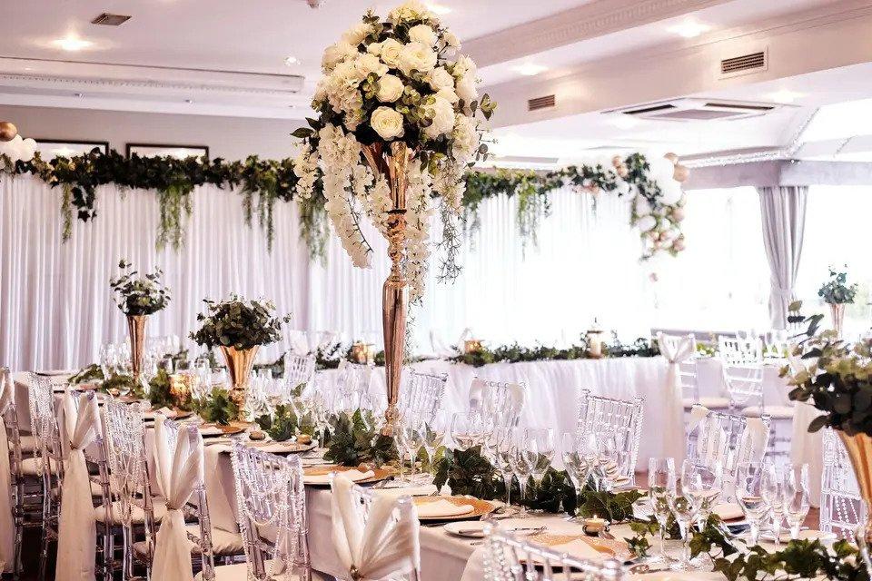 White reception room with long tables, chairs, chair sashes, foliage and white floral centrepieces 