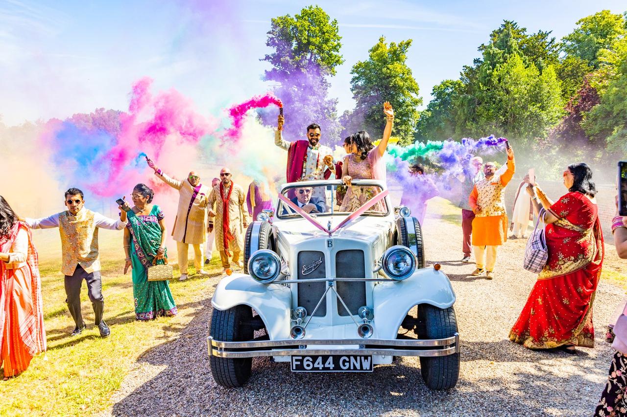 Indian bride and groom riding in a vintage wedding car surrounded by rainbow smoke and their friends
