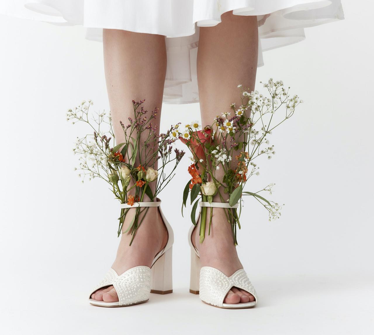 Close up of a model wearing a pair of pearl-embellished sandals. She has stems of flowers stuffed into the ankle straps.