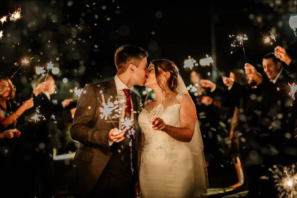 Bride and groom kiss outdoors while holding sparklers surrounded by guests holding sparklers