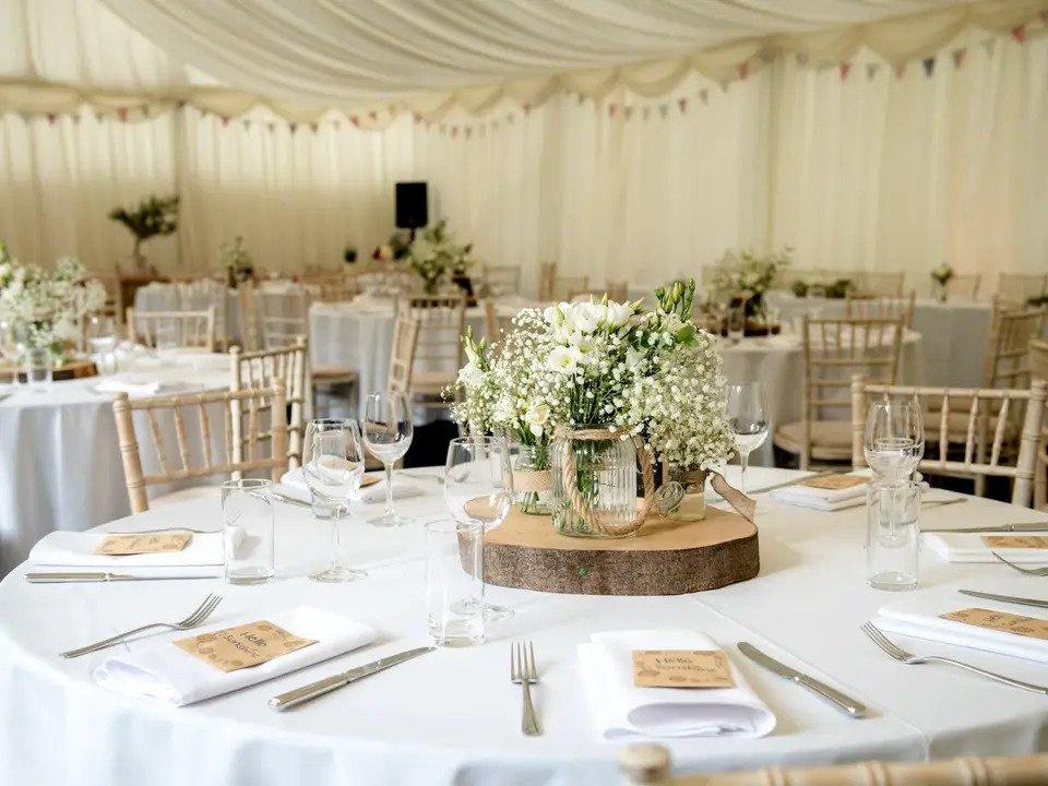 Close up shot of table in a marquee set for a wedding meal with white round tables, white Chiavari chairs, bunting, speaker set up and wooden and floral centrepieces