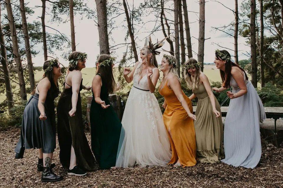 Bride and bridesmaids dancing together outdoors wearing headdresses made from leaves and greenery, and mismatched bridesmaid dresses in varying shades