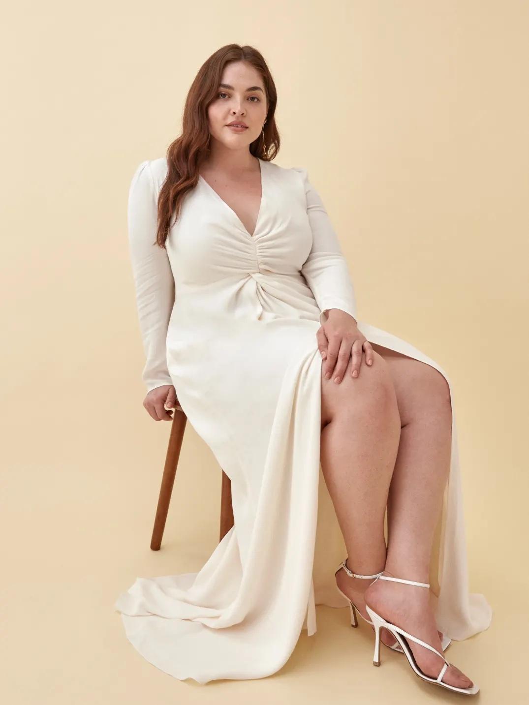 White woman with auburn hair sitting on a stool wearing a long sleeved white wedding dress with a ruched V neckline and split skirt