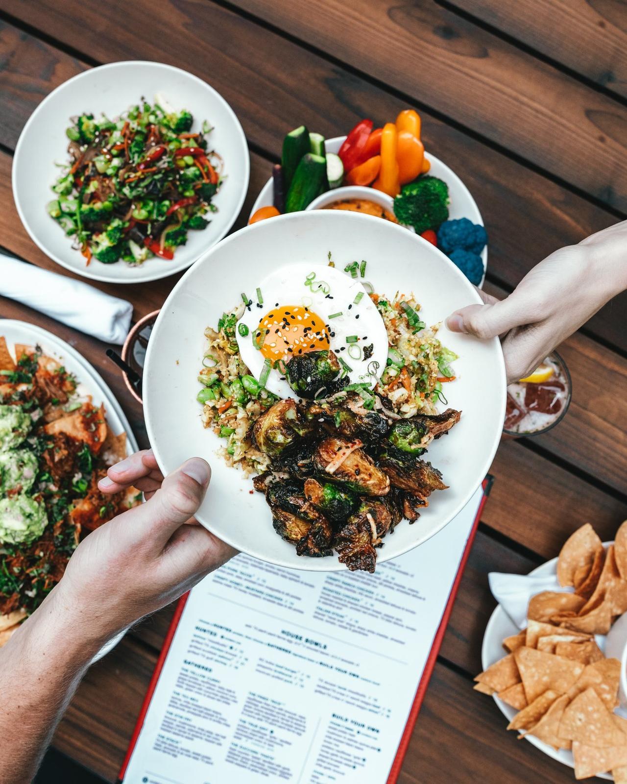 Couple reaching over a table each holding an Asian dish with fried egg and spring onions and vegetables with a spread of vegetable dishes below on a wooden table