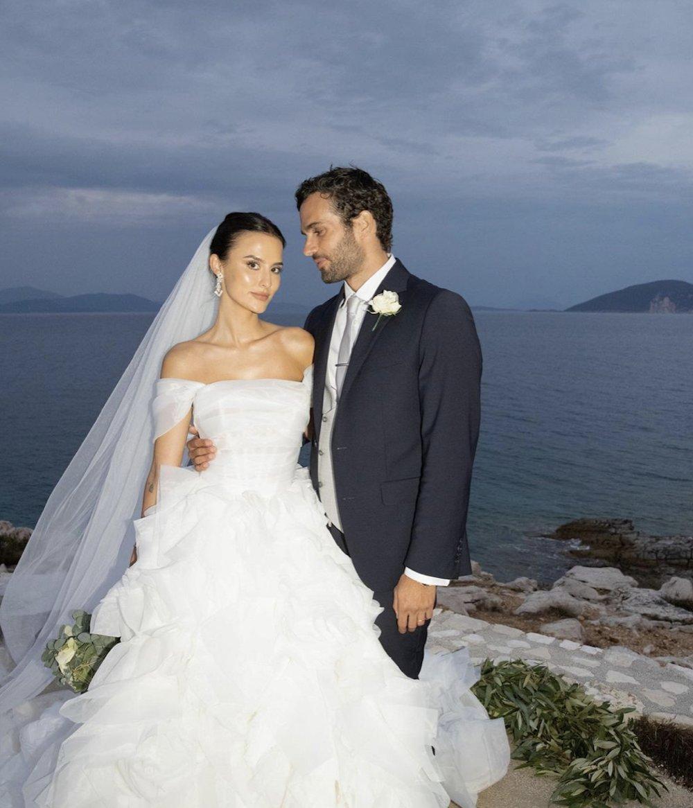 Lucy Watson on a Greek clifftop with her husband James Dunmore with Lucy in an off the shoulder tiered skirt white wedding dress and long sheer veil with drop diamond earrings and James in a dark grey suit