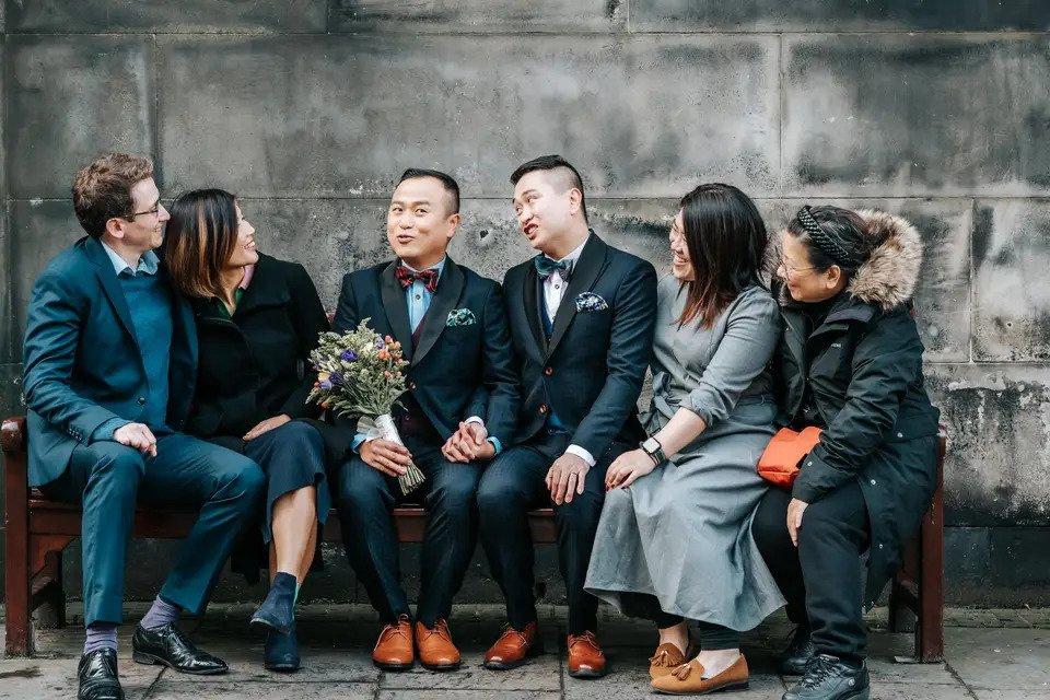 Male same sex couple sitting on a bench holding hands whilst surrounded by family members and friends. One of the grooms holds a bouquet of flowers