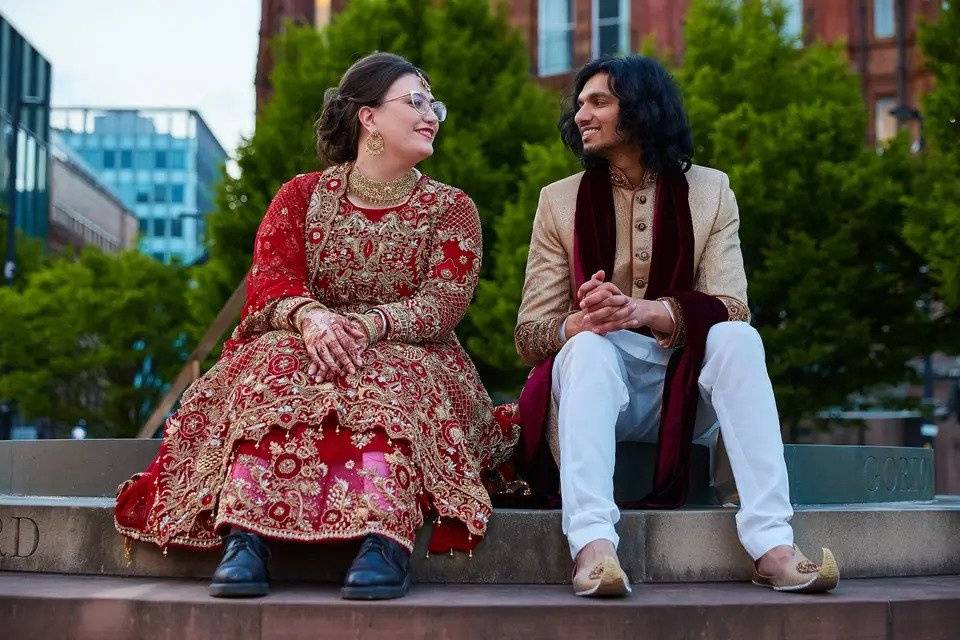 A couple in traditional Indian wedding attire sit on steps together smiling