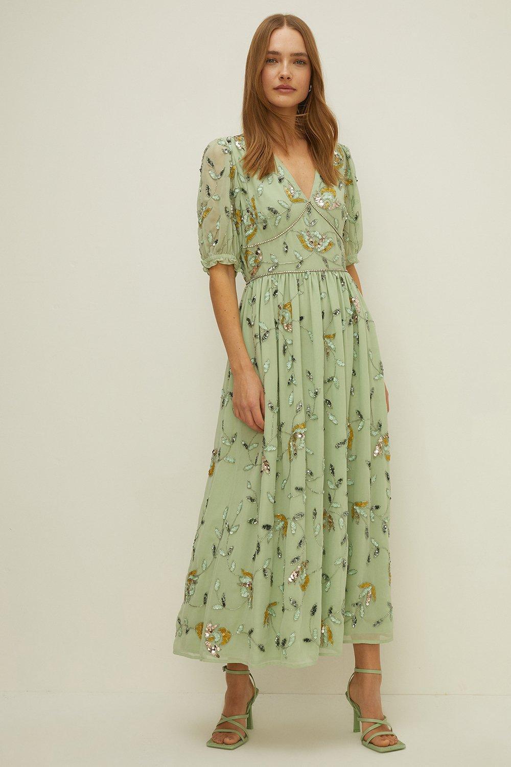 Model wearing green maxi dress with sleeves and v neck, covered in sequins and beaded embellishments