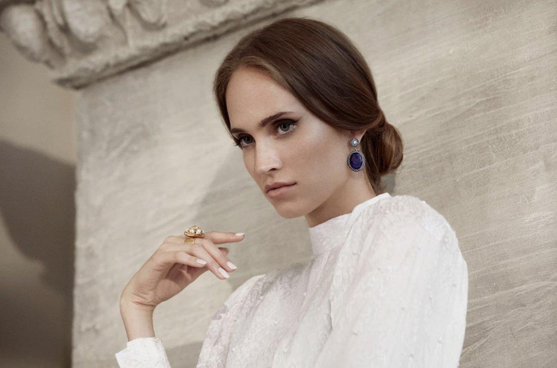 White woman with light brown hair wearing a high necked white dress, low bun, chunky gold ring and large oval sapphire earrings