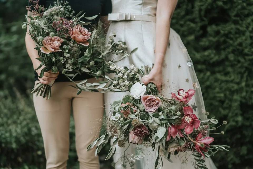 Bride in a dress next to a person in a gender neutral wedding outfit of trousers and a top - both clutch matching bouquets and only the bottom halves of their bodies are visible