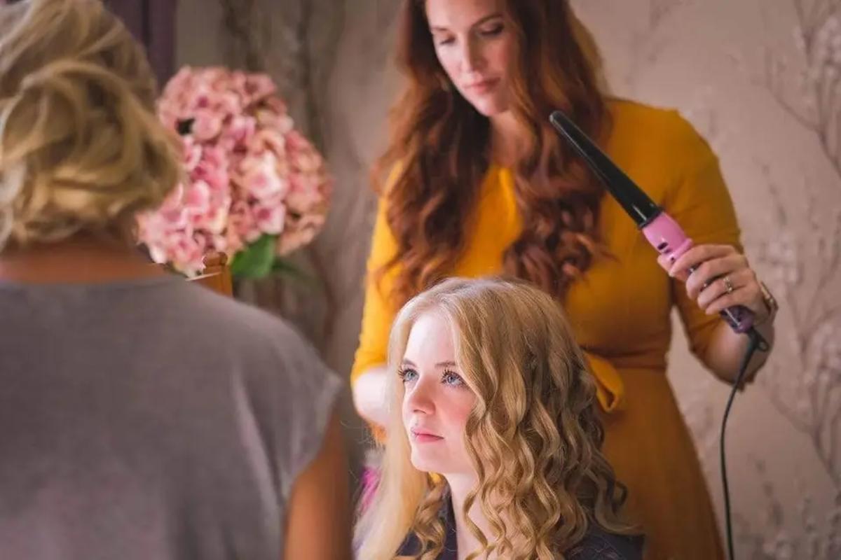 a hair and makeup artist half way through curling a blonde bride's medium length hair on the morning of her wedding