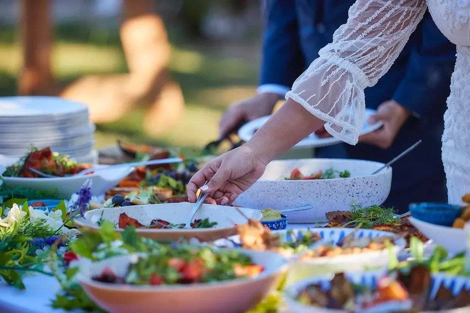 An outstretched hand serves salad from one of several salad bowls on a table decorated with fresh flowers