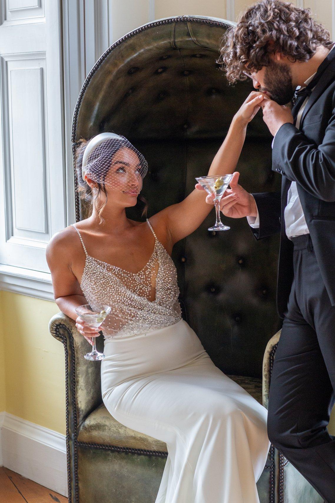 A groom kissing a bride on the hand as they sip martinis